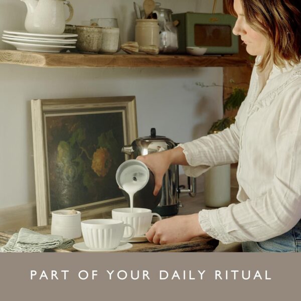 Woman using Dualit Handheld Milk Frother to pour frothed milk into a cup in a cozy kitchen.